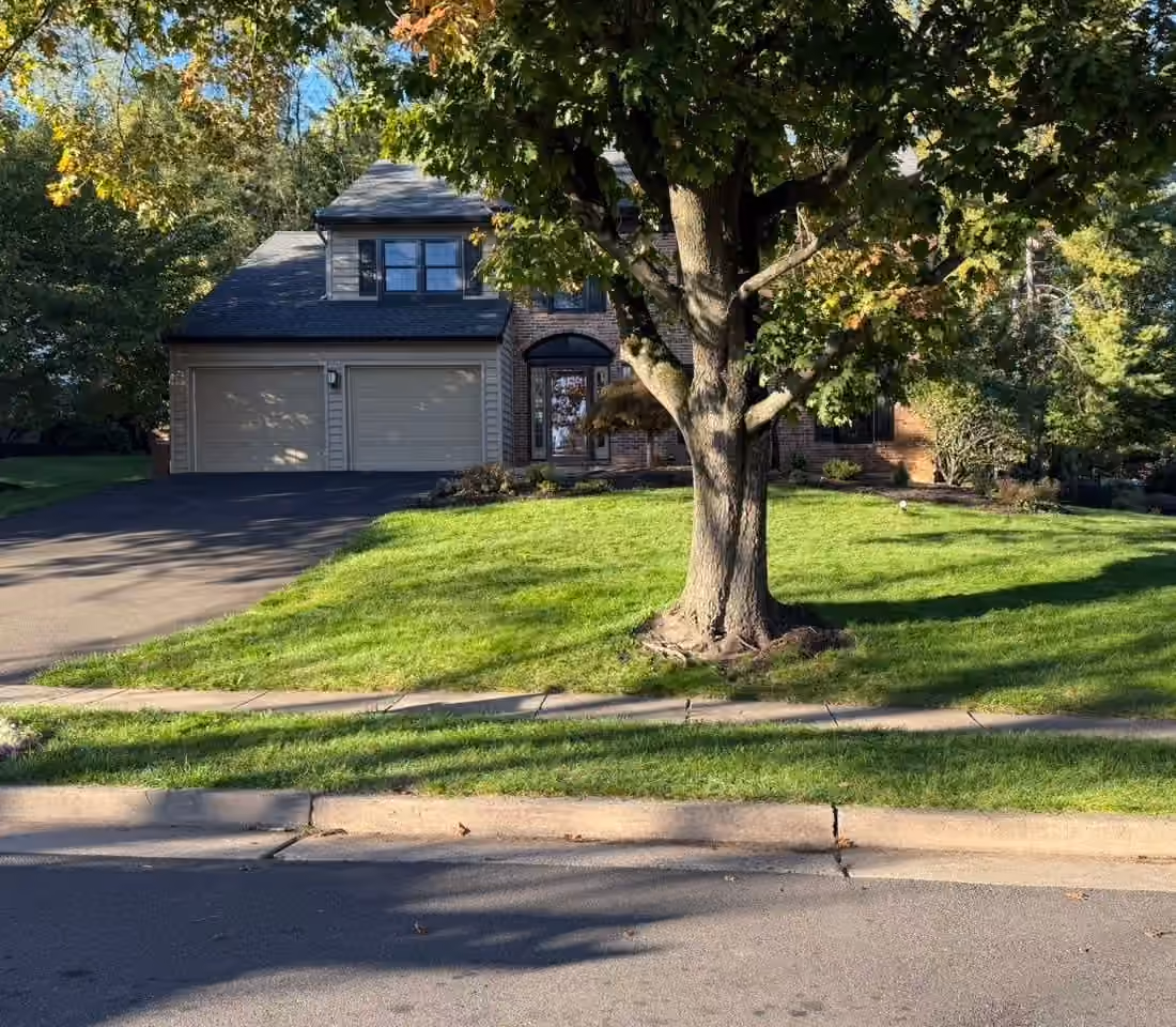 The same sloped front lawn clear of leaves, the brick house and oak behind it
