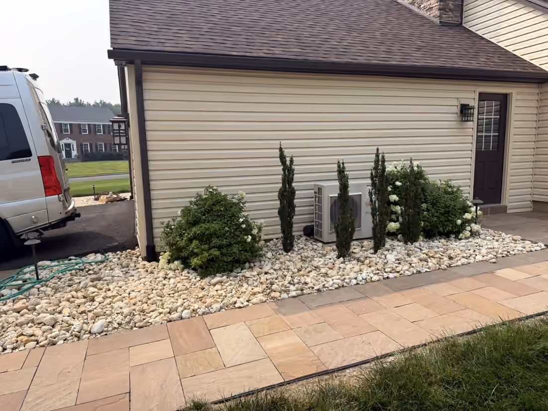 Upright evergreens and a hydrangea in a river rock bed beside a paver patio