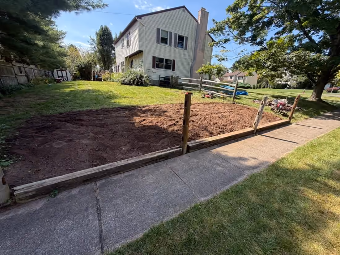 Wide patch of raked bare soil behind timber edging beside the sidewalk and the side of a house