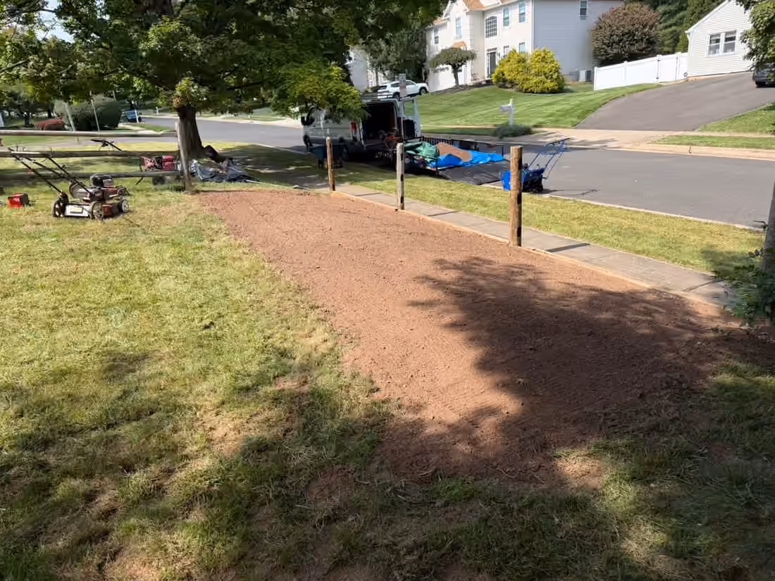 Long strip of freshly raked soil between a lawn and the sidewalk, with mowers and a loaded trailer at the curb
